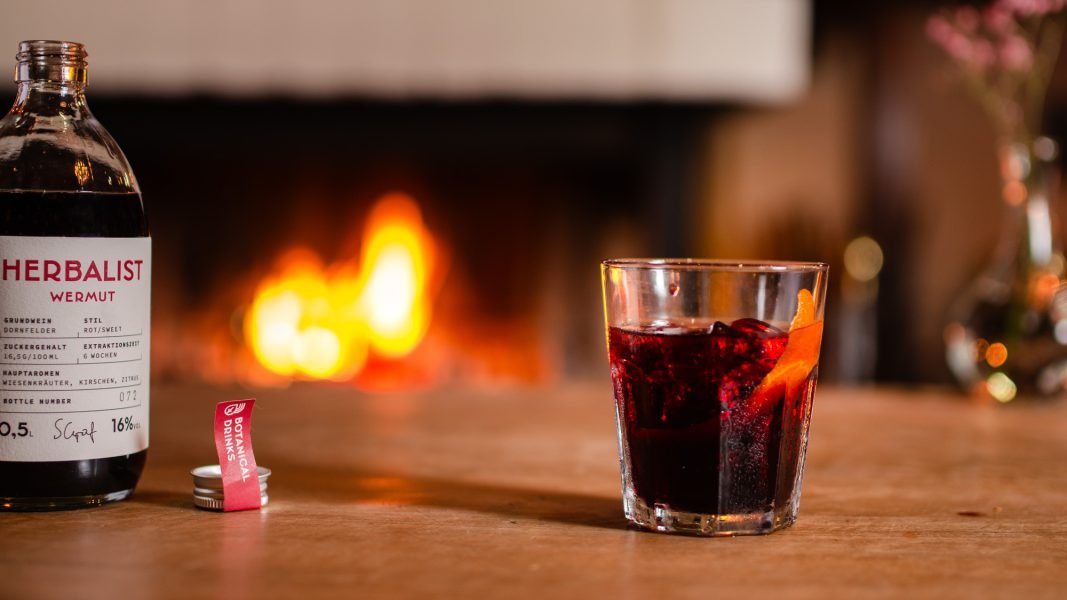 Herbalist Rot – Kaminmoment mit Drink und Flasche Glas mit rotem Wermut und Orangenzeste neben einer „Herbalist“-Flasche, auf Holztisch vor einem prasselnden Kaminfeuer